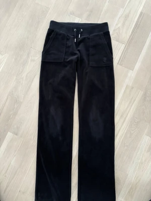 Svarta velourbyxor Juicy Couture XS - Svarta velourbyxor från Juicy Couture i storlek XS. Byxorna har bred resårmidja med snörning, stora fickor fram och bak med lock och silverfärgade knappar. Mjukt material och klassisk straight passform, perfekt för chill och streetstyle. En knapp saknas på fickan bak, annars fint skick.