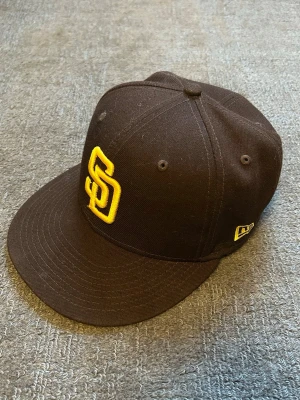 Brun San Diego Padres keps New Era - Säljer en brun San Diego Padres 59FIFTY keps från New Era med gul broderad SD-logga framtill och MLB-logga bak. Kepsen är tillverkad i 100% polyester och har en klassisk, rak skärm. Perfekt för dig som gillar baseball eller vill ha en snygg streetstyle-accessoar.