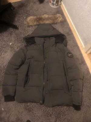 Svart pufferjacka från Canada Goose  - Canada Goose pufferjacka i svart med huva och avtagbar fuskpäls. Jackan har dragkedja och knappar framtill, två sidofickor och ribbade muddar vid ärmslut. Märkespatch på ärmen och quiltad design för extra värme. Perfekt för kalla vinterdagar.