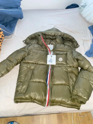 Olivgrön Moncler pufferjacka med huva - Säljer en olivgrön pufferjacka från Moncler med huva och ikonisk logga på bröstet. Jackan har en snygg röd, vit och blå dragkedja, praktiska fickor och quiltad design. Perfekt för kalla dagar och riktigt stilren. Byten kan godkännas, pris kan även det diskuteras.