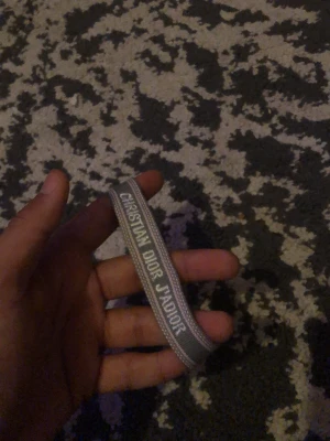 Grå Christian Dior J'Adior armband - Snyggt grått armband från Christian Dior med texten 'J'Adior' broderad i vitt. Armbandet har en enkel och stilren design med tunna broderade kanter och är gjort i ett mjukt tygmaterial. Perfekt accessoar för att lyfta din outfit.