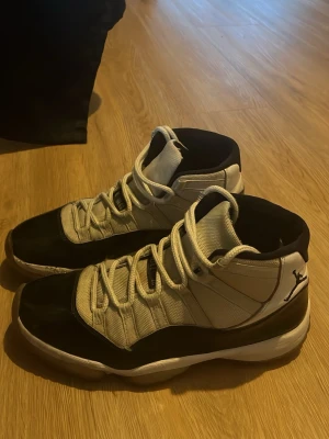 Nike Jordan 11 svart och vit sneakers - Nike Jordan 11 sneakers i svart och vit med högt skaft och snörning. Skorna har en rund tå, platt sula och tydlig Jumpman-logga på hälen samt siffran 45. Ovandelen är i syntet och skinn, med blank finish på den svarta delen. Perfekt för dig som gillar klassiska basketmodeller.