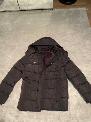 Svart pufferjacka med huva - Säljer en svart pufferjacka med huva och mjukt foder på insidan. Jackan har dragkedja och knappar framtill, samt flera fickor med knappstängning. Perfekt för kalla dagar och har en schysst, quiltad look som är både snygg och praktisk.