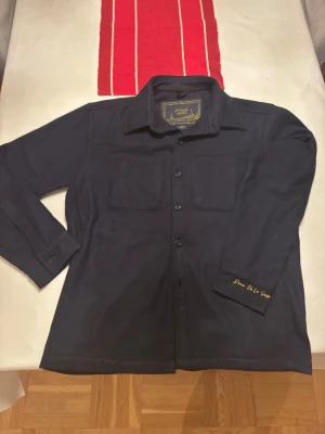  overshirt från D'once De La Sense - Mörkblå overshirt i ull från D'once De La Sense, med klassisk krage, två bröstfickor och knäppning framtill. Snygg broderad logga i gult på ärmen ger en unik touch. Perfekt för lager-på-lager och stilrena outfits. Knapped längst ner gick sönder när jag provade den men det går enkelt att laga med reservknappen