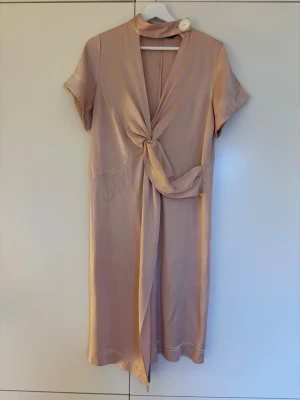 Gammelrosa omlottklänning med drapering från Zara, storlek S - Snygg gammelrosa klänning i glansigt viskosmaterial med korta ärmar och drapering framtill. Klänningen har en hög krage med dekorativ knapp och djup v-ringning. Materialet känns lyxigt och har en mjuk satinliknande finish. Midilängd. Jättefint skick! Storlek S