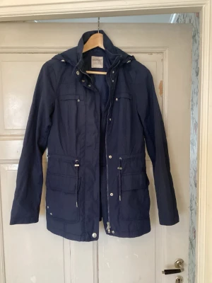 Marinblå parkajacka från Esprit - Snygg marinblå parkajacka från Esprit med dragsko i midjan, flera praktiska fickor och huva. Jackan har dragkedja och knappar framtill och är perfekt för dig som vill ha en stilren och funktionell jacka till vardagen.