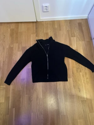 Svart stickad kofta med dragkedja 157 - Svart stickad kofta från 157 i storlek S. Modellen har en hög krage och hel dragkedja framtill. Materialet är grovstickat och ger en schysst struktur. Perfekt för lager-på-lager och chill dagar. Långa ärmar och klassisk passform.