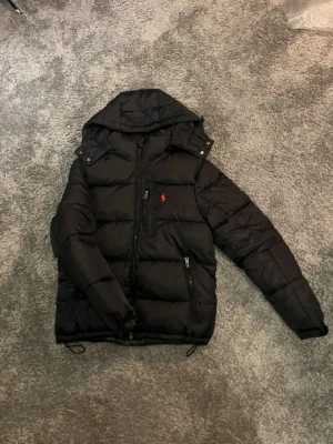 Svart pufferjacka från Polo Ralph Lauren - Svart pufferjacka med huva från Polo Ralph Lauren. Jackan har flera dragkedjeförsedda fickor, quiltad design och en röd Polo-logga på bröstet. Materialet är polyester och jackan har långärmade ärmar samt snygga detaljer med knappar vid huvan.