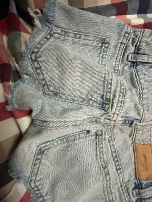 Ljusblå jeansshorts från Gina Tricot - Säljer ett par ljusblå jeansshorts från Gina Tricot med fransiga kanter och klassisk femficksdesign. Shortsen har bälteshällor, knapp och dragkedja framtill. Perfekta för varma dagar och har en avslappnad vibe.