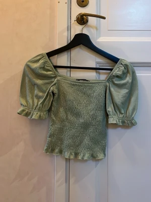 Grön blus med puffärm från Gina Tricot - Säljer en ljusgrön blus från Gina Tricot i storlek XS. Toppen har puffiga korta ärmar med volangkant och smockad kropp som ger en snygg och bekväm passform. Perfekt för vår och sommar, och den har en söt och feminin stil.