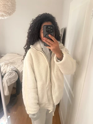 Vit teddyjacka med dragkedja - Vit teddyjacka med dragkedja framtill och två stora fickor. Jackan har quiltat foder på insidan för extra värme samt lite oversized passform. Den är använd två gånger och köpt för 700kr. Perfekt som vinterjacka eller även till våren. Passar både M och S
