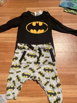 Batman set från DC Comics barnkläder - Säljer ett Batman-set från DC Comics med långärmad svart tröja och gråa byxor, båda med det klassiska Batman-loggan i gult och svart. Byxorna har snörning i midjan och mjuk resår. Perfekt för alla små superhjältar!