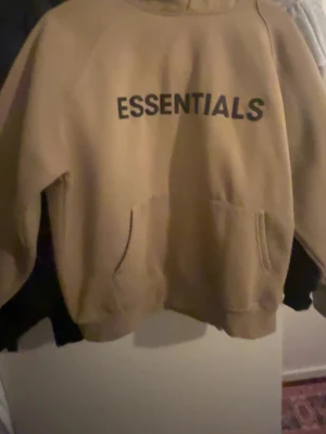 Essentials - Beige hoodie från Essentials i mjukt, kraftigt material. Stilren design med diskret logotyp framtill. Bekväm passform med huva och magficka. Perfekt för vardag, streetwear eller lager-på-lager. Använd men i fint skick, utan större slitage. Ett säkert basplagg. Passar både dam och herr, lätt att matcha året runt i garderoben enkelt.
