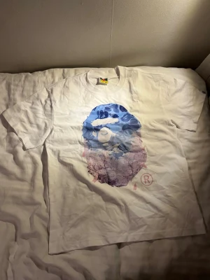 Vit bathing ape bape t-shirt med blått tryck - Säljer en vit t-shirt från BAPE i storlek M med ikoniskt ape-tryck på bröstet. Trycket har blå och lila toner med kamouflagemönster och naturmotiv. Klassisk rund hals och korta ärmar. Perfekt för dig som gillar streetwear och vill sticka ut.