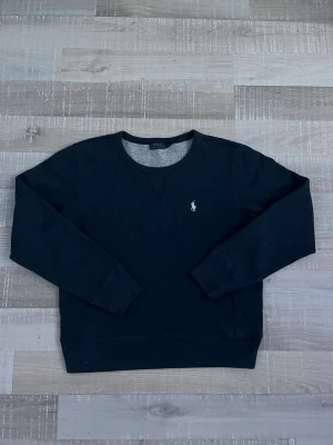 Mörkgrå sweatshirt från Polo Ralph Lauren - Mörkgrå sweatshirt från Polo Ralph Lauren med klassisk rund halsringning och broderad vit logga på bröstet. Tröjan är i mjuk bomulls- och polyestermix och har långa ärmar samt ribbade muddar vid ärmslut och nederkant. 