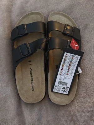 Svarta sandaler från Daily Essentials - Säljer ett par svarta sandaler från Daily Essentials med två breda remmar och justerbara spännen. Innersulan är beige och har en mjuk yta som känns skön mot foten. Perfekt för varma dagar och enkel att matcha med olika outfits.