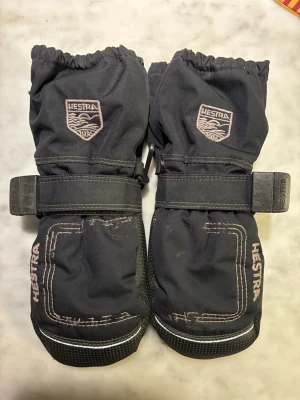 Svarta Hestra tumvantar med dragkedja - Säljer ett par svarta tumvantar från Hestra med justerbart band runt handleden och dragkedja på sidan. Vantarna har broderad logga och detaljer i vitt samt greppvänlig prickstruktur i handflatan. Perfekta för kalla vinterdagar.