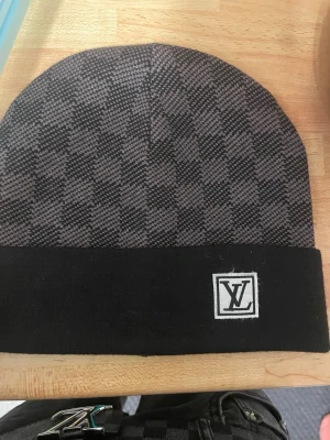 Rutig svart väst från Louis Vuitton - Snygg väst från Louis Vuitton i svart och grått med rutig design. Västen har en klassisk form och är tillverkad i ett mjukt material som känns lyxigt. LV-logga framtill ger en exklusiv touch. Perfekt för dig som gillar designerplagg och vill sticka ut.