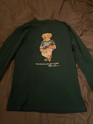 Mörkgrön långärmad Polo Bear tröja barnstorlek XL - Säljer en mörkgrön långärmad tröja från Ralph Lauren med ikoniskt Polo Bear-tryck på bröstet. Tröjan är i mjuk bomull och har en klassisk rund hals. Perfekt för dig som gillar streetwear och vill sticka ut med en cool print.