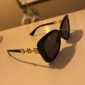 Svarta solglasögon med pärldetaljer - Snygga svarta solglasögon med breda bågar i fyrkantig form. Sidorna har lyxiga guldfärgade detaljer med pärlor och små stenar som ger en exklusiv vibe. Perfekt accessoar för att höja din stil och sticka ut i mängden.