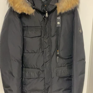 Svart dunjacka med päls från Hollies - Svart dunjacka från Hollies med snygg päls på huvan och flera praktiska fickor framtill. Jackan har lång ärm, dragkedja och knappar, samt en cool badge på ärmen. Perfekt för kalla dagar och har en riktigt schysst streetstil.