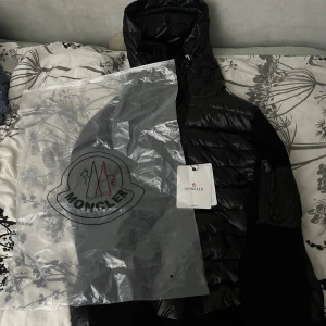 Svart Moncler pufferjacka med huva - Svart pufferjacka från Moncler med glansig yta och stickade svarta ärmar. Jackan har huva, dragkedja framtill och en ficka med dragkedja på ärmen. Ikonisk Moncler-logga på ärmen och medföljande originalpåse. Perfekt för kalla dagar. Helt Nytt, aldrig använd, perfekt för vintern ❄️