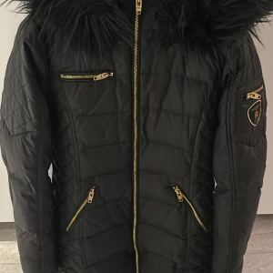 Svart Rockandblue dunjacka med fuskpäls - Snygg svart dunjacka från Rockandblue med stor fluffig fuskpäls på huvan och guldfärgade dragkedjor. Jackan har quiltade partier på sidorna, flera fickor med dragkedja och Rockandblue-logga på ärmen. Perfekt för kalla vinterdagar.