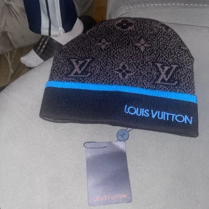 Louis Vuitton mössa med blå kant - Snygg svart och mörkbrun mössa från Louis Vuitton med klassiskt monogrammönster och en blå kant. LV-loggan är broderad i blått framtill. Mössan är stickad och har en exklusiv känsla, perfekt för dig som vill ha något extra till vintern.