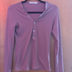 ribbad långärmad tröja NA-KD - Snygg långärmad tröja från NA-KD i en mjuk rosa ton. Tröjan har ribbad struktur, v-ringning och silverfärgade knappar framtill. Perfekt för dig som gillar enkel men stilren design med lite edge.