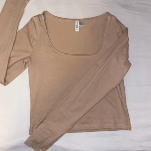 Beige långärmad topp från H&M - En stilren beige långärmad topp från H&M divided med fyrkantig ringning. Toppen är lite croppad och har en enkel, clean design som passar perfekt till jeans eller kjol. Tillverkad i mjuk bomull för en skön känsla hela dagen.