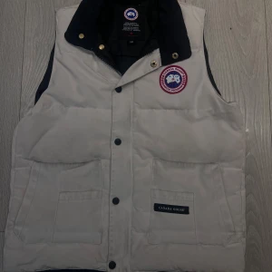 Vit dunväst från Canada Goose - Snygg vit dunväst från Canada Goose med klassisk logga på bröstet och svarta detaljer vid kragen. Västen har två stora fickor framtill och stängs med knappar. Perfekt för lager-på-lager och riktigt clean look.