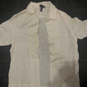 Vit kortärmad skjorta från H&M - Snygg vit skjorta från H&M med korta ärmar och krage. Skjortan har ett diskret prickigt mönster och knappar framtill. Materialet är lätt och känns som bomull, perfekt för en chill look. Passformen är normal och skjortan har en klassisk form.