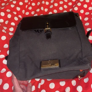 Svart ryggsäck från Gaston Luga - Snygg svart ryggsäck från Gaston Luga med detaljer i skinn och guldfärgade spännen. Väskan har ett lock med spänne, justerbara axelremmar och en stilren metallbricka framtill. Insidan är fodrad i vinrött tyg med logotyp. Perfekt för dig som vill ha en stilren och praktisk väska.nypris 1000kr.