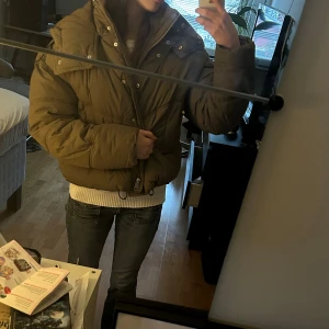 Kahki grön pufferjacka  - Säljer en kahki grön lite croppad pufferjacka från H&M i storlek S. Jackan har en stor, mysig huva, dragkedja och knappar framtill samt quiltade detaljer. Passformen är loose och jackan är perfekt för kalla dagar. Skriv vid frågor! 