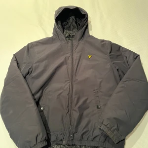 Svart vindjacka från Lyle & Scott - Svart vindjacka från Lyle & Scott med huva och gul logotyp på bröstet. Jackan har dragkedja framtill, två fickor med tryckknappar och elastiska muddar vid ärmslut och nederkant. Perfekt för kyliga dagar och enkel att matcha med jeans eller hoodie.