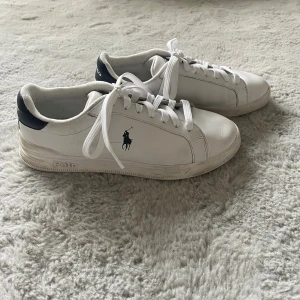 Vita sneakers från Polo Ralph Lauren - Snygga vita sneakers från Polo Ralph Lauren med marinblå detaljer på hälen och klassisk Polo-logga på sidan. Jag har använt dessa skor ganska mycket men det är fortfarande i bra skick!