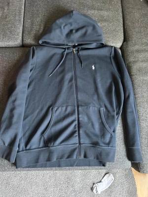 Mörkblå hoodie från Polo Ralph Lauren XL - Snygg mörkblå hoodie från Polo Ralph Lauren i storlek XL. Tröjan har huva med snörning, dragkedja framtill och två rymliga fickor. Klassisk Polo-logga broderad på bröstet. Tillverkad i mjuk bomull för skön komfort. 