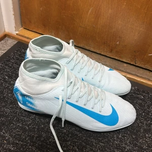 Nike Mercurial vita fotbollsskor - Nike Mercurial fotbollsskor i ljusblått och vitt med högt skaft och snörning. Snyggt blått Nike-märke på sidan och Mercurial-tryck baktill. Skorna har platt sula med dobbar för gräsplan och är tillverkade i syntetmaterial för lätt känsla och bra grepp.