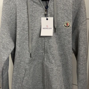Grå Moncler hoodie med dragkedja - Snygg grå hoodie från Moncler med dragkedja framtill och klassisk logga på bröstet. Tröjan har huva med snörning och två fickor. Perfekt för chill dagar och enkel att matcha med jeans eller joggers. Tveka inte att komma med prisförslag!!