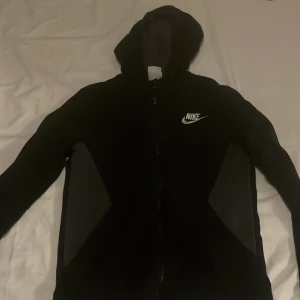 Svart hoodiejacka från Nike - Svart hoodiejacka från Nike med vit logga på bröstet. Jackan har hel dragkedja, huva och två sidofickor med mörkgrå paneler. Tillverkad i mjuk bomullsmix, perfekt för chill eller träning. Snygg och enkel design som funkar till det mesta.