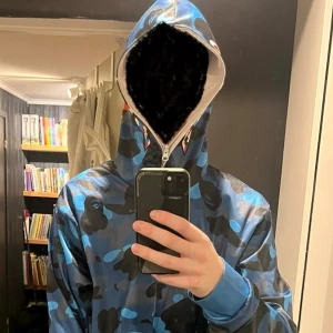 BAPE blå camo shark hoodie - Säljer en BAPE shark hoodie i blått camo-mönster med ikonisk hajdesign på huvan och broderade bokstäver. Tröjan har hel dragkedja som går upp över ansiktet, ribbade muddar och detaljerade tryck på ärmarna. Perfekt för dig som vill sticka ut med streetwear.