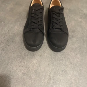 Svarta sneakers från Christian Louboutin - Snygga svarta sneakers från Christian Louboutin i mocka med klassisk rund tå och platt sula. Skorna har svarta snören och den ikoniska röda undersidan med Louboutin-logga. Insidan är beige med guldfärgad branding. Perfekt för dig som gillar exklusiv streetstyle.