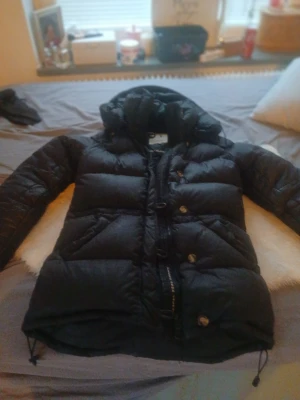 Svart dunjacka med huva från boomarang  - Säljer en svart, fluffig dunjacka från Downemong med huva och snörning nertill. Jackan har både dragkedja och knappar framtill, samt breda quiltade sektioner som ger en cool och chunky look. Perfekt för kalla dagar när du vill hålla värmen och se chill ut.