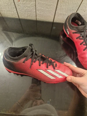 Röd/rosa  adidas fotbollsskor strl 38 - Säljer ett par röd rosa adidas fotbollsskor med svarta snören och vita klassiska ränder på sidorna. Skorna har plös i svart och mönstrad ovandel i syntetmaterial. Yttersulan har fasta dobbar för bra grepp på planen. Perfekta för dig som vill sticka ut på matchen!