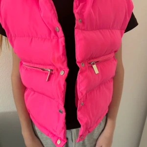 Neonrosa dunväst med dragkedjor - Snygg neonrosa dunväst med puffig look och två fickor med silvriga dragkedjor framtill. Västen har tryckknappar längs hela framsidan och en cool, sportig vibe. Perfekt för att sticka ut och hålla värmen under kyliga dagar.