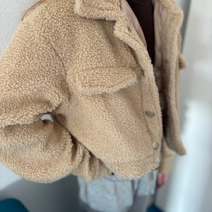 Beige teddyjacka med broderi - Säljer en beige teddyjacka från Hot Damn med broderad röd text på bröstet. Jackan har stora fickor med lock, krage och knäppning med silvriga knappar. Den är croppad och har långärmade ärmar, perfekt för att ge en chill vibe till din outfit.