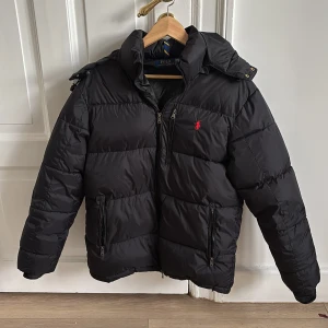 Ralph Lauren pufferjacka - Svart pufferjacka från Polo Ralph Lauren med röd logga, perfekt nu för vinter då kyla inte är något problem du kommer dras med. Jackan är inköpt för mer än 1 år sen på zalando men är fortfarande i grymt skick utan några hål eller andra defekter. Hör av er vid intresse💸🕺