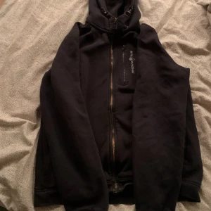 Svart hoodie från Sail Racing - Svart hoodie från Sail Racing med dragkedja framtill och huva. Tröjan har en bröstficka med dragkedja och Sail Racing-logga på bröstet. Perfekt för kyliga dagar och enkel att matcha med det mesta.