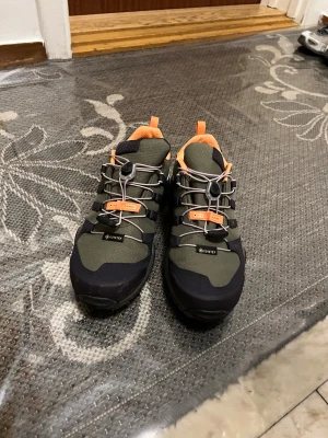 Adidas goretex  - Adidas sneakers i mörkgrönt och svart med orange och vita detaljer. Skorna har snabbsnörning, platt sula och rund tå. Tillverkade i syntetmaterial med Gore-Tex för vattentäthet. Perfekta för utomhusaktiviteter och äventyr. Användes en tillfälle