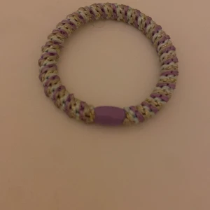 Färgglatt flätat armband i lila och beige - Säljer ett snyggt armband med flätad design i lila, beige och inslag av guld. Armbandet har en rund form och en lila detalj som accent. Perfekt accessoar för att lyfta din stil.